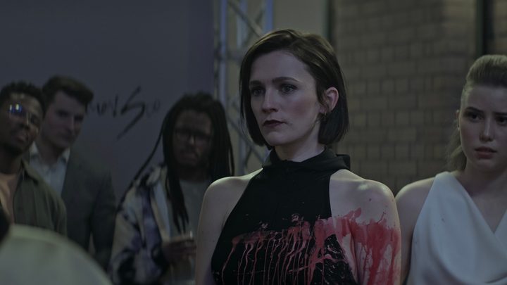 Kate from You season 4 looking upset, wearing a black halter dress that is covered in red paint.