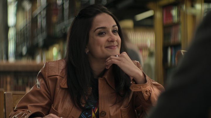 Nadia from You season 4 sitting at a table in a library, hand on her chin, smiling.