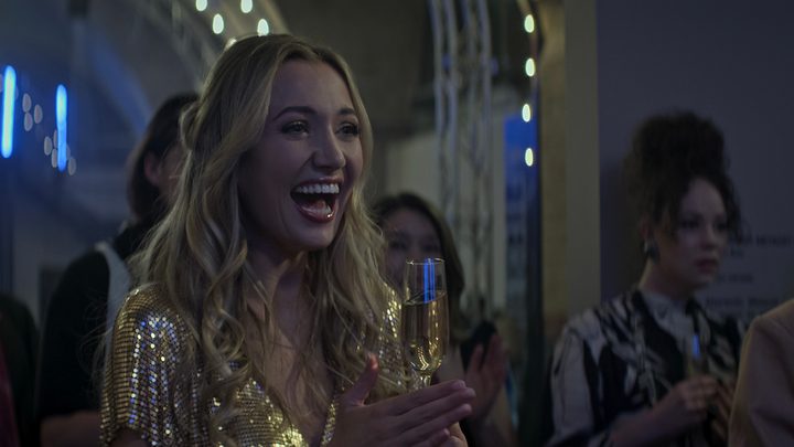 Lady Phoebe from You season 4 at a party, laughing and holding a glass of champagne.