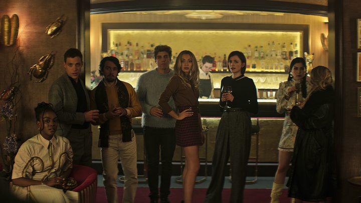 The new characters on You season 4 standing at a bar, dressed to the nines, looking at something.