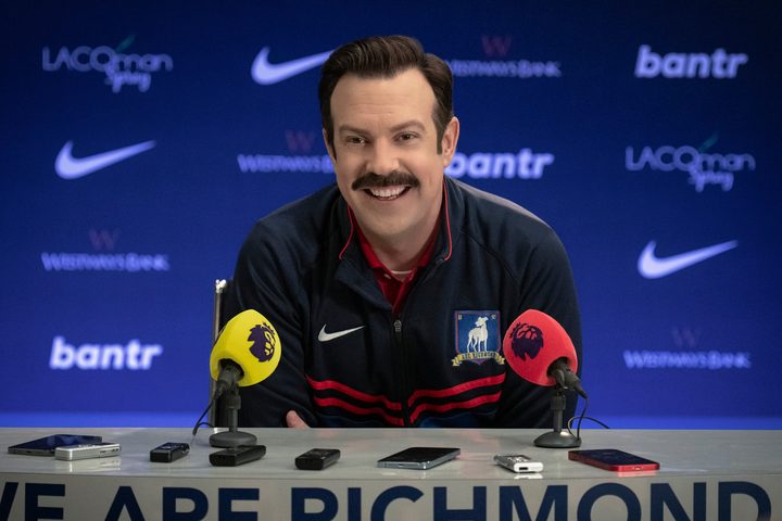 Ted Lasso sits at a press conference table in season 3.