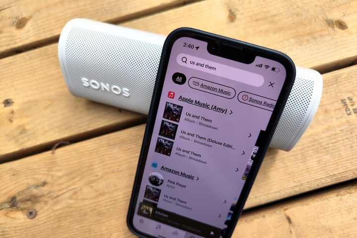 An iPhone 14 displaying the Sonos app search feature, sitting in front of a Sonos Roam speaker.