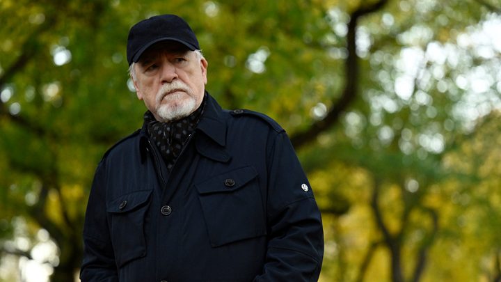 Logan wearing a black baseball hat and jacket, standing outside looking straight ahead in Succession.