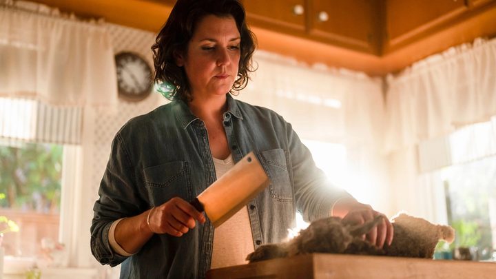 Shauna from Yellowjackets cooking dinner in the kitchen, holding a big cleaver to cut meat and looking emotionless.