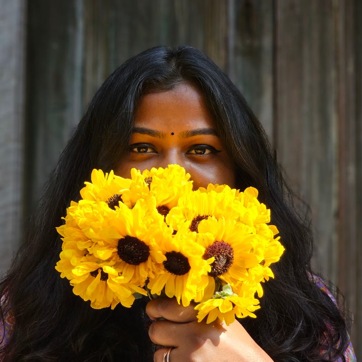 Woman looks at camera while holding a bunch of sunflowers in her hand captured with the Samsung Galaxy S23 Ultra’s 3x telephoto portrait mode camera.