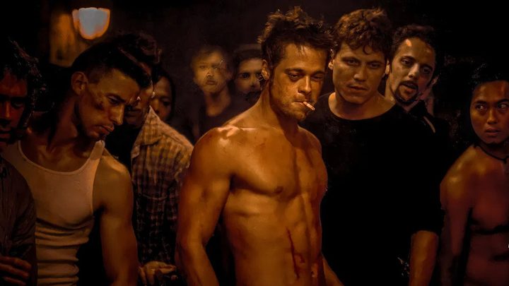 Tyler Durden from Fight Club standing shirtless, cigarette in his mouth looking down with a group of other men behind him.
