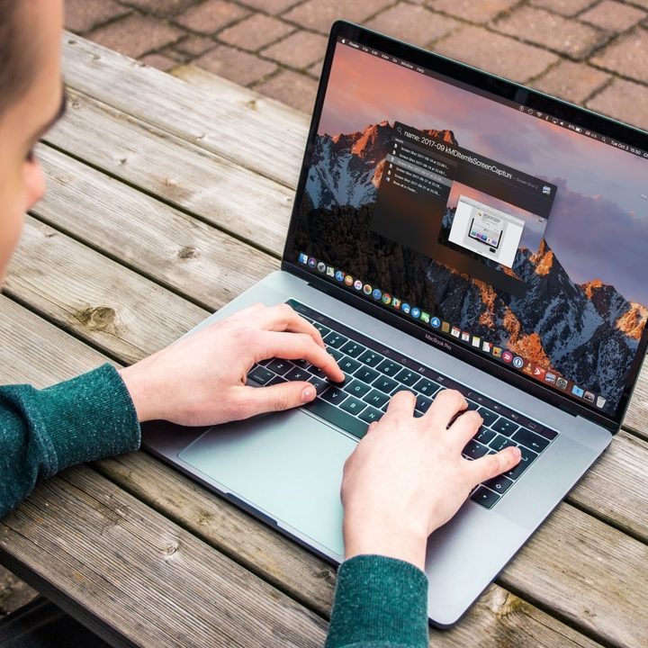 A person typing on a MacBook Pro while sat on a wooden bench.