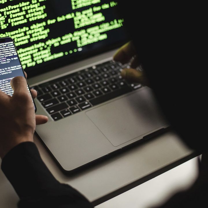 A hacker typing on an Apple MacBook laptop while holding a phone. Both devices show code on their screens.