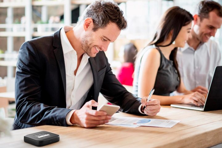 Man working on a laptop and holding a phone in public area with a mobile hotspot on the table.