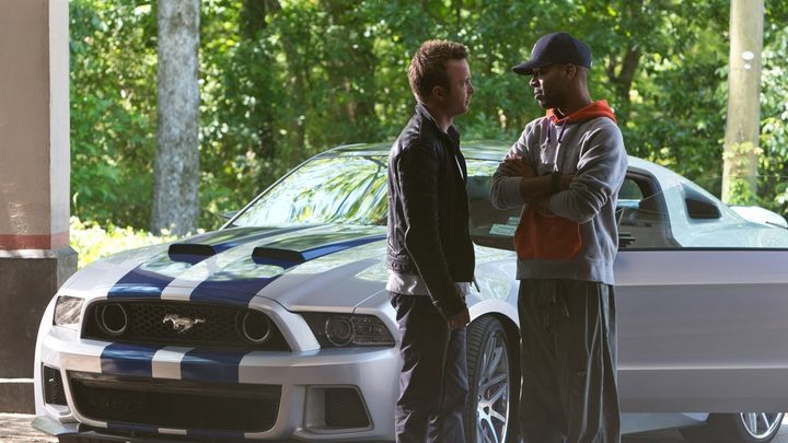 Two men talk in front of a car in The Need for Speed.