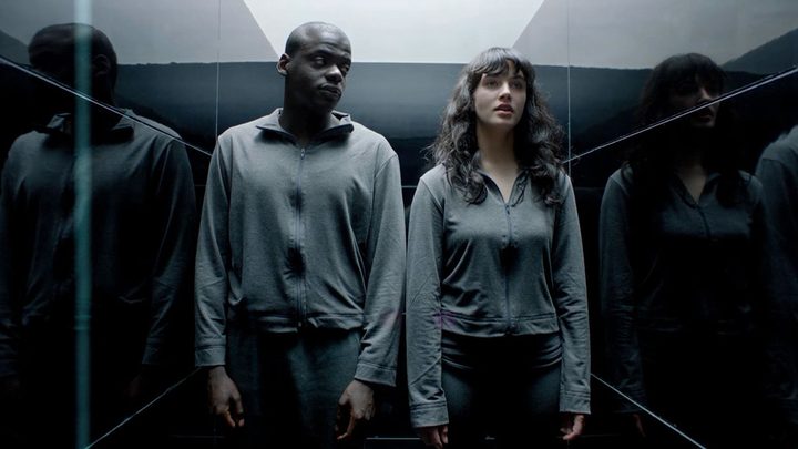 A young man and woman stand side by side in an elevator, both wearing drab grey sweaters in a scene from the Fifteen Million Merits episode of Black Mirror.