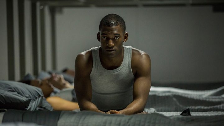 Stripe from the Black Mirror episode Men Against Fire sitting on his bed in a tank top looking worried.