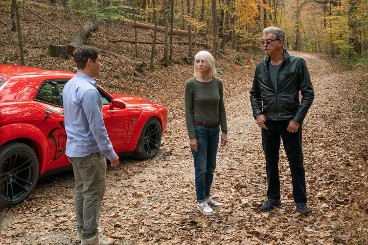 Adam Devine, Ellen Barkin, and Pierce Brosnan stand near a red muscle car in The Out-Laws.