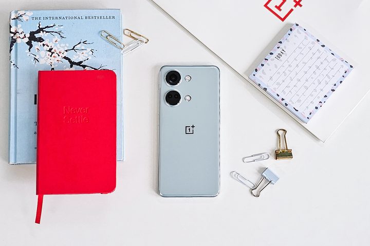 OnePlus Nord Misty Green color on white background with a red Never Settle notebook, post it notes, clips, and a blue book on the side.