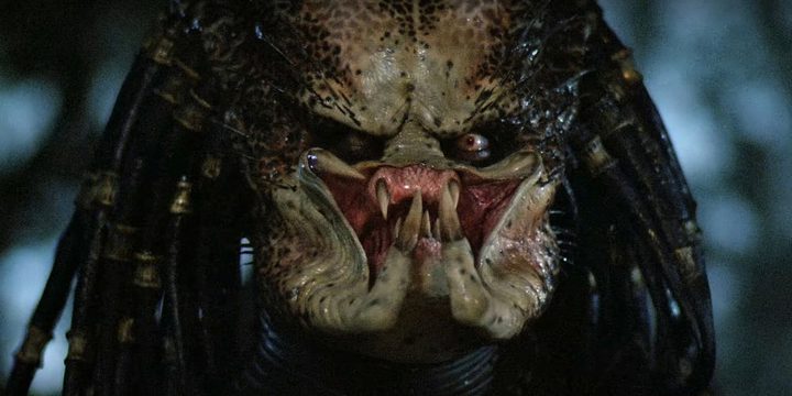 The Predator takes off his mask, revealing his fangs.