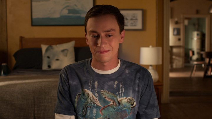 A young man sits in his room looking down and smiling in a scene from Atypical.