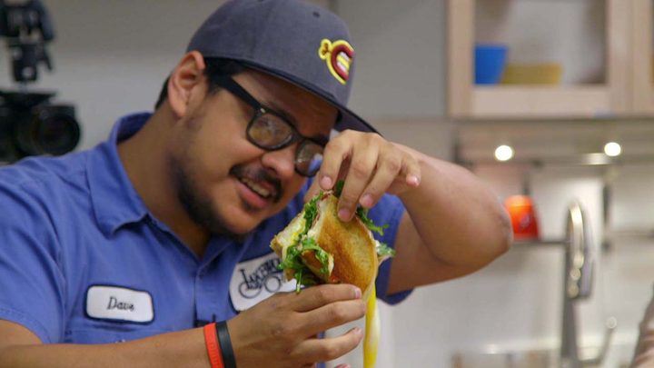 A man holding a sandwich in the air looking at it, cheese oozing out in a scene from Cooking on High.