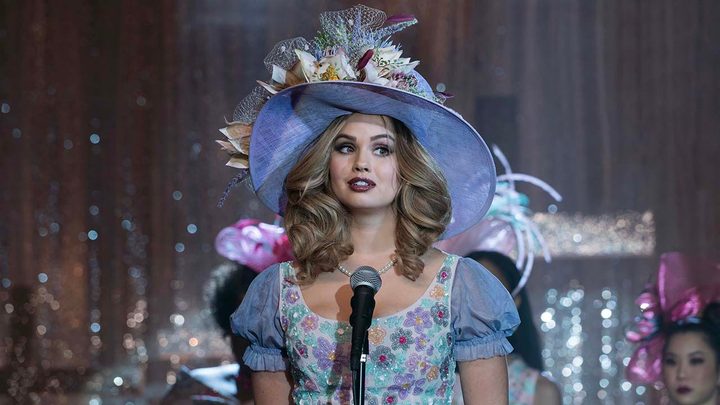 A young woman wearing an oversized, wide-brimmed hat and blue dress standing in front of a microphone in a scene from Insatiable.