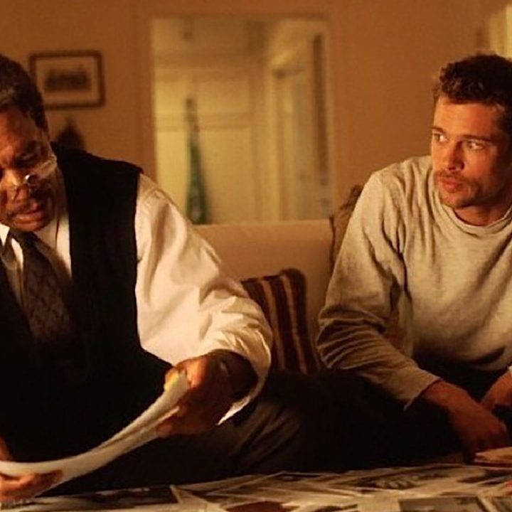 Morgan Freeman and Brad Pitt sit together on a couch looking at papers in a scene from Seven.