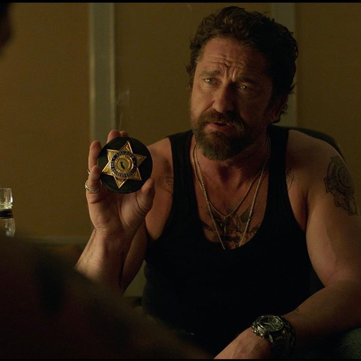 Gerard Butler sits down and holds up a police badge in Den of Thieves.