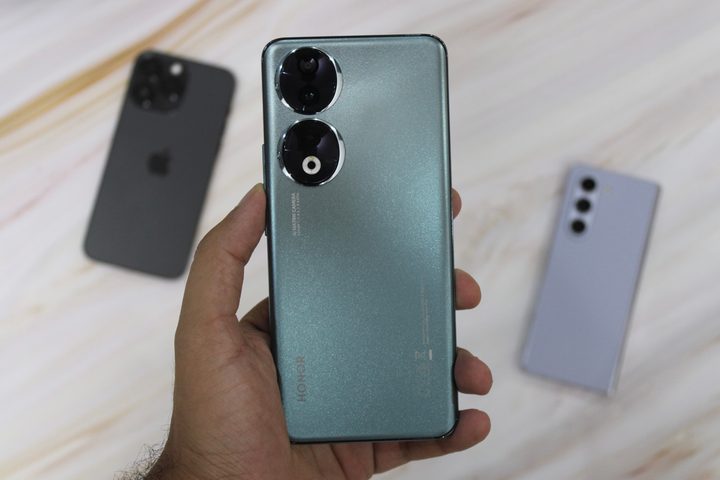 Honor 90 in hand with iPhone 14 Pro Max and Galaxy Z Fold 5 in background.