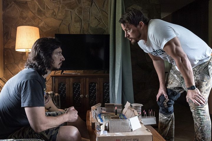 Adam Driver sits on the couch as Channing Tatum stands over him in Logan Lucky.