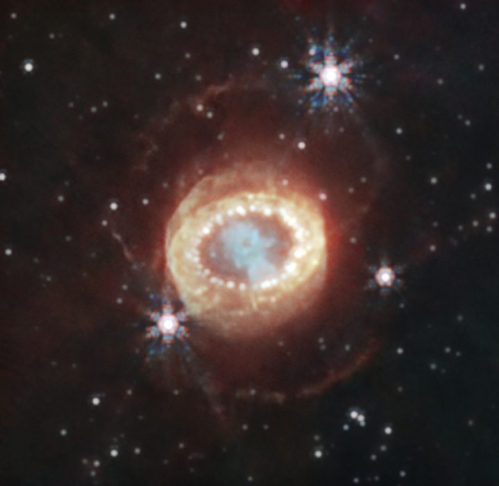Webb’s NIRCam (Near-Infrared Camera) captured this detailed image of SN 1987A (Supernova 1987A). At the center, material ejected from the supernova forms a keyhole shape. Just to its left and right are faint crescents newly discovered by Webb. Beyond them an equatorial ring, formed from material ejected tens of thousands of years before the supernova explosion, contains bright hot spots. Exterior to that is diffuse emission and two faint outer rings. In this image blue represents light at 1.5 microns (F150W), cyan 1.64 and 2.0 microns (F164N, F200W), yellow 3.23 microns (F323N), orange 4.05 microns (F405N), and red 4.44 microns (F444W).