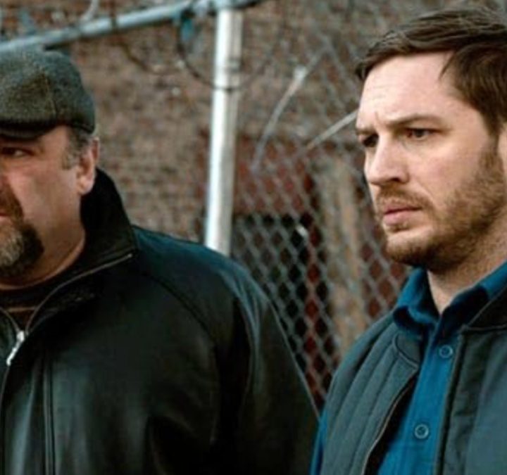 James Gandolfini and Tom Hardy stand next to each other in front of a fence in The Drop.