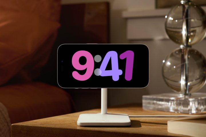 iPhone on charging stand showing floating clock in iOS 17 StandBy mode.
