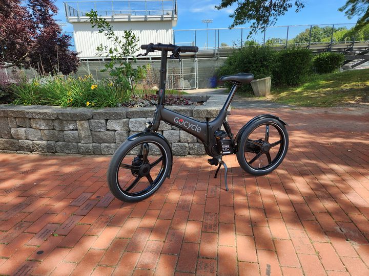 A front three-quarter view of the left side of a GoCycle G4 folding e-bike parked on a high school campus with an athletic stadium in the background.