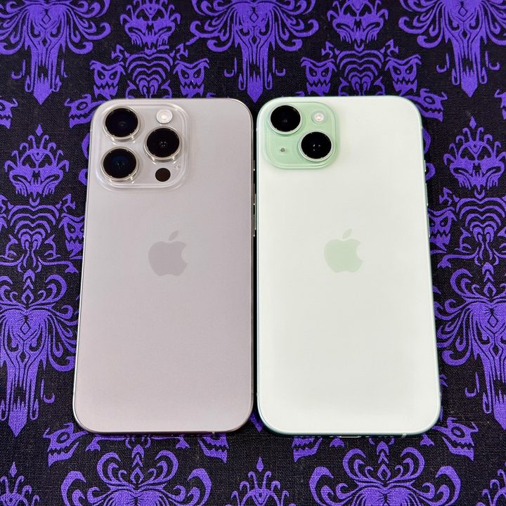 A natural titanium iPhone 15 Pro (left) and a green iPhone 15 on a Haunted Mansion wallpaper placemat.