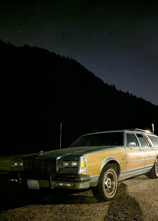A wood paneled Buick Lesabre in front of a pitch black mountain, with stars shining in the background