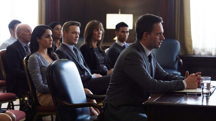 Mike sitting in court in a scene from Suits, people sitting behind him watching the trial.