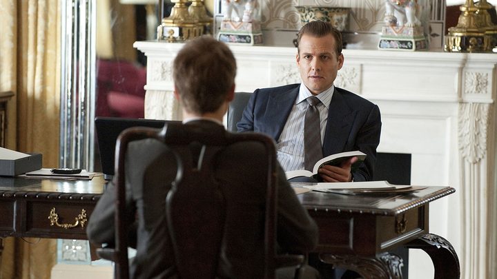 Harvey sitting at his desk talking to Mike who is facing him in a scene from Suits.