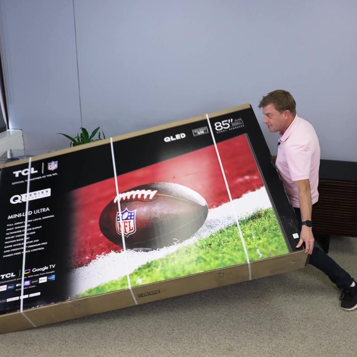 A man struggles to put an 85-inch boxed TV upright in a studio space.