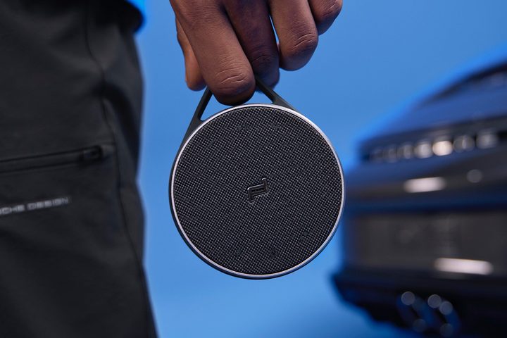 Hand carrying Porsche Design PDS20 Bluetooth speaker in front of a Porsche car.