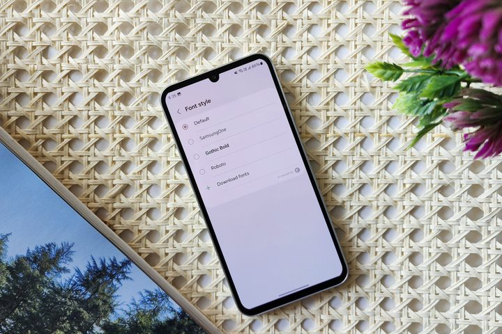Samsung One UI 6 Sans font on Galaxy A34 kept beside purple flowers and cooffee table book on a retan surface table.