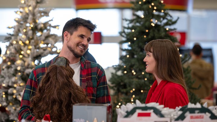 Robbie Amell and Leighton Meester in a festive store looking at one another, smiling in a scene from the movie EXmas.