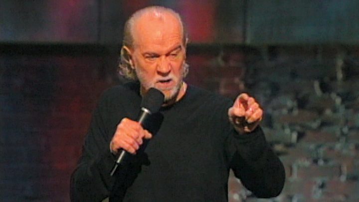 George Carlin on stage with a mic pointing a finger and wincing his face in a scene from George Carlin: You Are All Diseased.