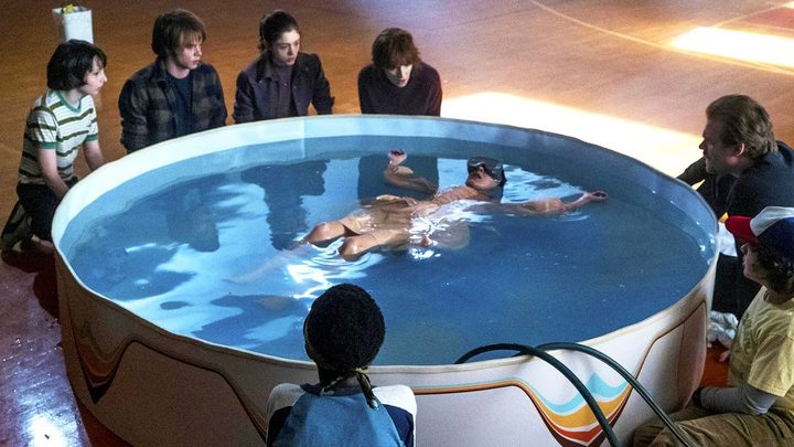 Eleven lying in a kiddie pool filled with water and Epsom salts while the other surround her in a scene from Stranger Things.