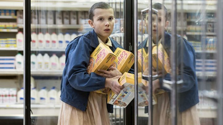Eleven standing in a grocery store freezer section clutching boxes of Eggs waffles in a scene from Stranger Things.