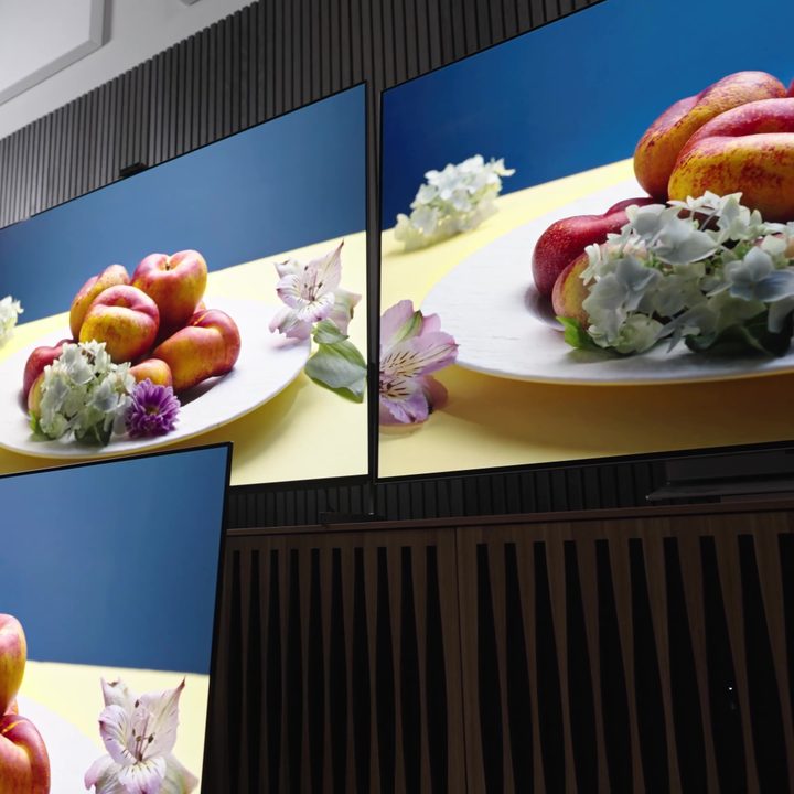 A platter of flowers and fruit against a blue background compared on a Samsung S95C, Sony A95L, LG G3, and Sonly A95K TV.