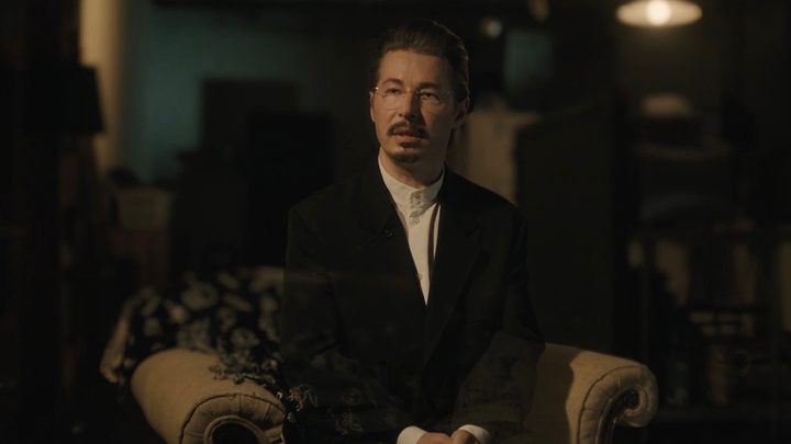 Ray in a suit sitting in a chair in a scene from A Murder at the End of the World.