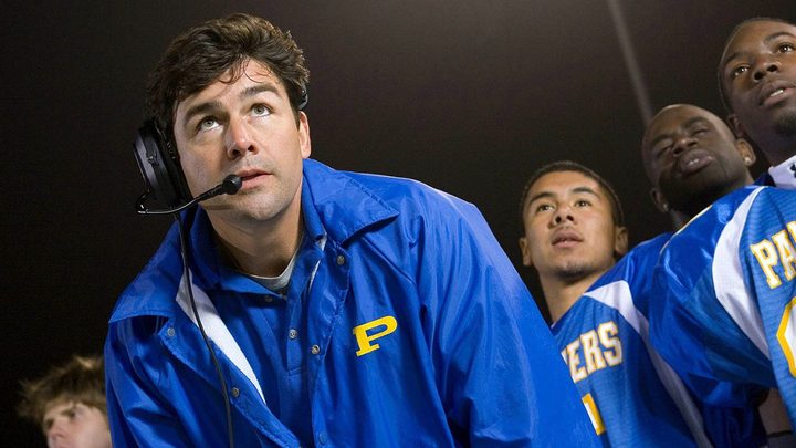 A man wearing a headset in a sports uniform, looking up from the sidelines in a scene from Friday Night Lights.