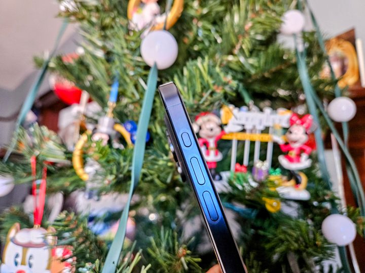 iPhone 15 Pro in Blue Titanium showing side view of Action and volume buttons in front of a Christmas tree.