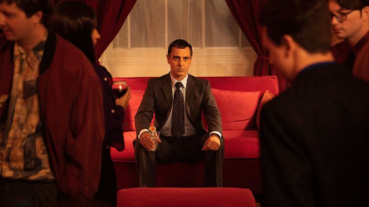 A man sitting in a suit in a red chair, others looking on in a scene from Sexy Beast on Paramount+.