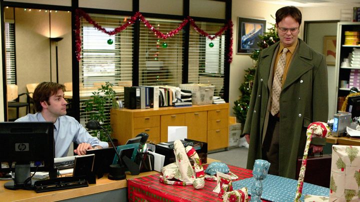 Two men sit and stand near a desk in The Office.