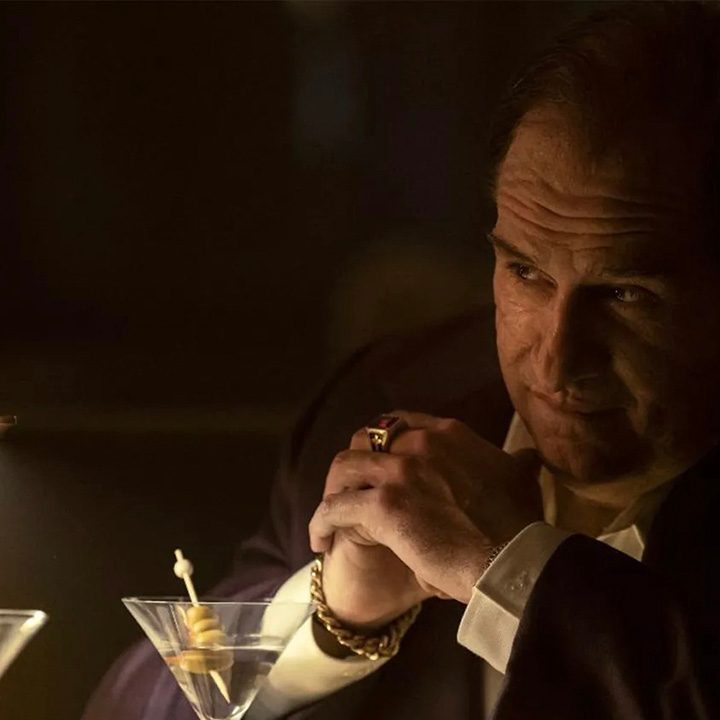 The Penguin sitting at a table in the dark, his hands clasped together in a scene from the series.