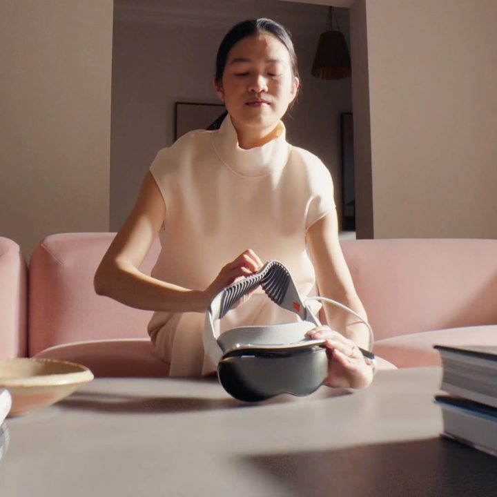A person picks up an Apple Vision Pro headset that is resting on a desk.