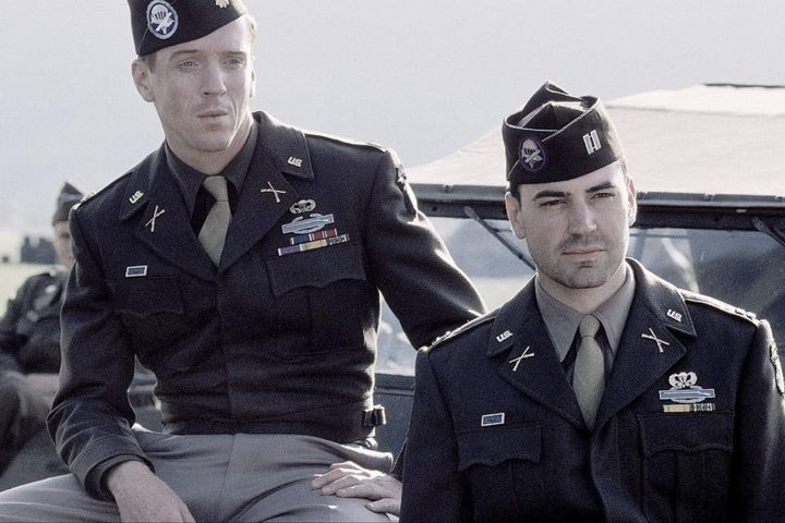 One soldier sits on a car while the other soldier stands next to him.